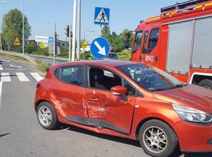 Czerwone renault clio po kolizji drogowej. bok samochodu uszkodzony. W tle straż pożarna, sygnalizacja świetlna i droga.