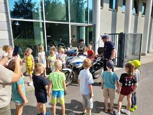 Policjant ruchu drogowego przy motocyklu policyjnym. Na motocyklu siedzi przedszkolak. Grupa dzieci gromadzi się przy motocyklu.