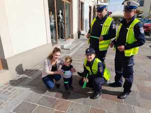 Policjanci ruchu drogowego i policjantka prewencji w odblaskowych kamizelkach. Policjantka kuca przy dziecku. Obok matka dziecka. W tle Rynek w Olkuszu i zabudowania.