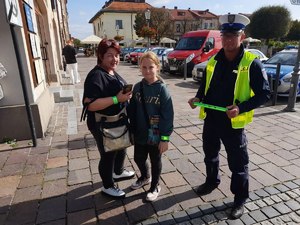 Policjantka w odblaskowej kamizelce wręcza przechodniom odblaski. W tle olkuski Rynek i zabudowania