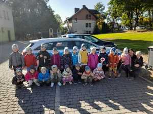 Przedszkolaki wraz z policjantka przy policyjnym radiowozie. Policjantka kuca przy przedszkolakach.  W tle budynki i parking.