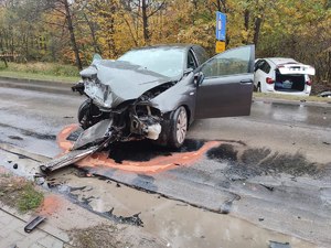 Samochody po kolizji. Przód czarnego auta zniszczony. Wgnieciona karoseria, rrozbita szyba, różne elementy na drodze. W tle las i biały samochód w rowie. Mokra nawierzchnia. Pora jesienna.