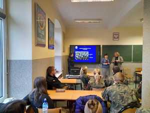 Policjantki z nauczycielem oraz rektorem uczelni podczas prelekcji. W tle prezentacja multimedialna. W ławkach szkolnych, na przeciwko prelegentów siedzą uczniowie w mundurach. Zdjęcie w sali lekcyjnej