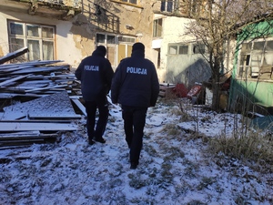 Dwaj policjanci przy pustostanie. Dookoła zniszczonego budynku panuje nieład (śmieci, stary desek) . Budynek zniszczony, wybite szyby w oknach.