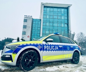 Nowo oznakowany radiowóz policyjny. W tle budynek Komendy Powiatowej Policji w Olkuszu. Pora zimowa, dookoła budynku i na jezdni śnieg.