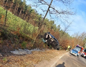 Wypadek drogowy. Samochód doszczętnie zniszczony. Auto w przydrożnym rowie. Na drodze radiowóz i wóz strażacki oraz strażacy. W tle las. Pogoda słoneczna, bezchmurne niebo.   (1)