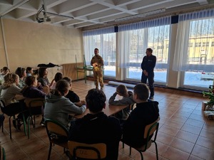 Strażak i policjantka podczas spotkania z dziećmi w klasie. dzieci siedzą w ławkach szkolnych. W tle okno i krajobraz.