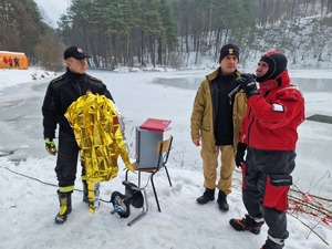 Ratownik WOPr oraz dwaj strażacy prowadza pogadankę. W tle zamarznięty staw. Krajobraz zimowy.