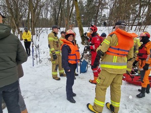 Policjantka w kamizelce ratunkowej o ochronnym kasku wśród grupy strażaków. W tle krajobraz zimowy i gromadzący się ludzie.