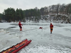 Dwóch strażaków w specjalnych ubraniach ratowniczych stoją w dużej odległości od siebie i za pomocą lin wydobywają poszkodowana osobę spod lodu. W tle zamarznięty staw i krajobraz
