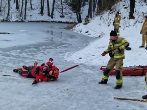 Strażak przy zamarzniętym stawie, ciągnie linę, przy której umocowany jest ratownik oraz poszkodowany. Symulacja ratowania człowieka spod lodu.