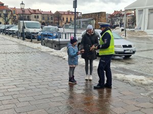 Policjantka ruchu drogowego (biała czapka) w kamizelce odblaskowej wręcza kobiecie i dziewczynce odblaski. W tle Rynek w Olkuszu oraz zaparkowane pojazdy. Zdjęcie zimą pogoda deszczowa.