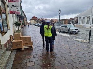 Policjantka ruchu drogowego w odblaskowej kamizelce wręcza przechodniom odblaski. W tle zaparkowane samochody, budynki i olkuski Rynek.